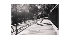 Attraversando il ponte, tra voce e silenzio Due bambini piccoli di spalle attraversano correndo un ponte di legno. La più grande stringe un peluche tra le braccia, davanti a sé, mentre dietro il più piccolo indossa un cappello.